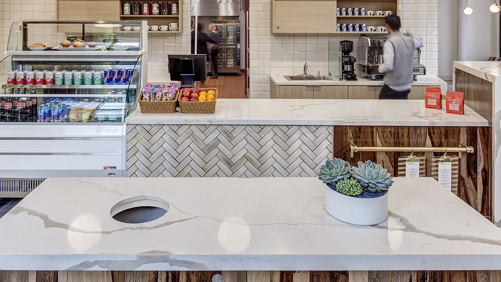 Modern cafe with white countertops and tiled walls, wood accents and flooring. A barista is making a drink behind the counter.